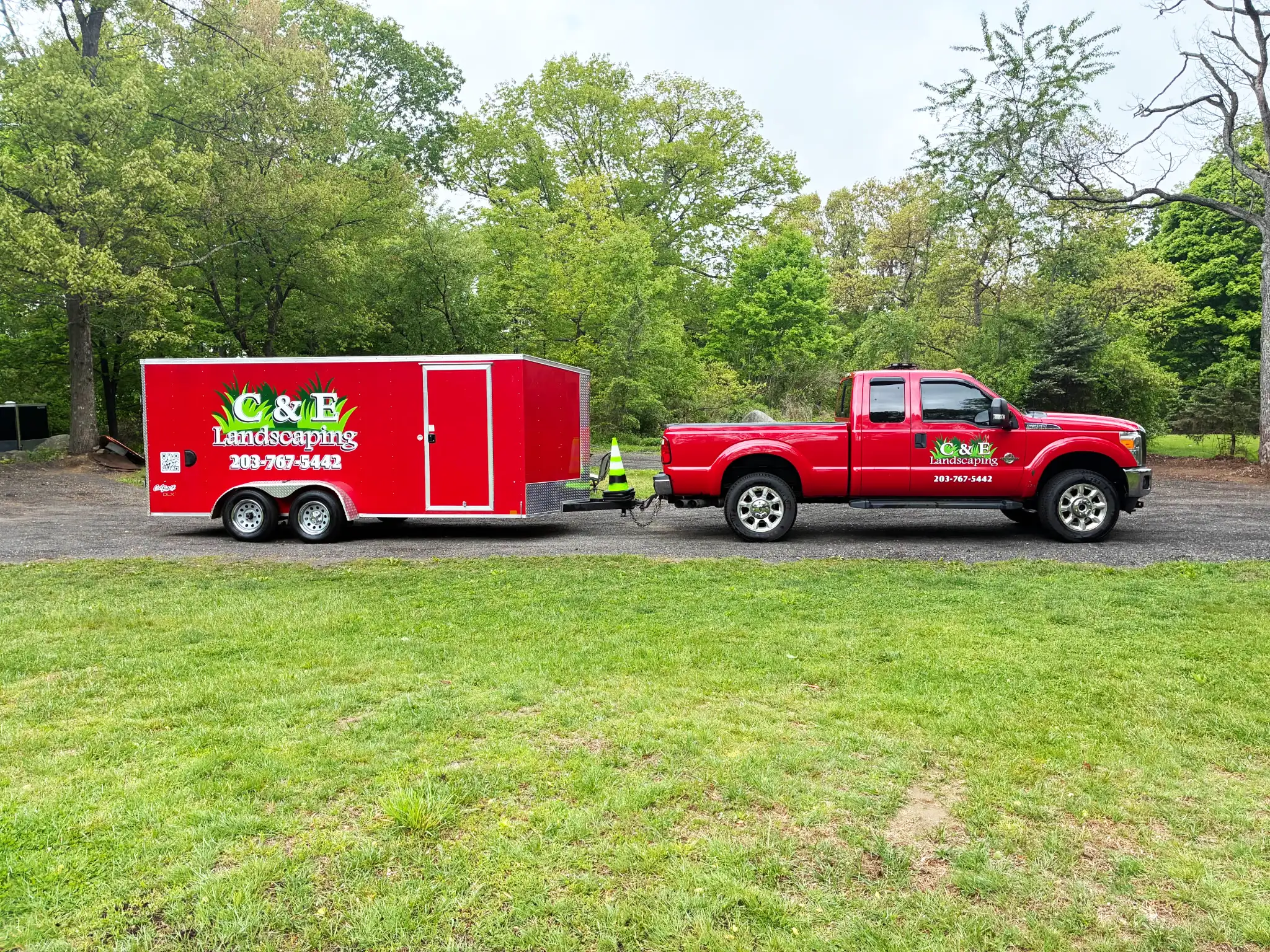 Red landscaping trailer hitched to a red pickup truck in a gravel lot, with green trees in the background and branding on both vehicles